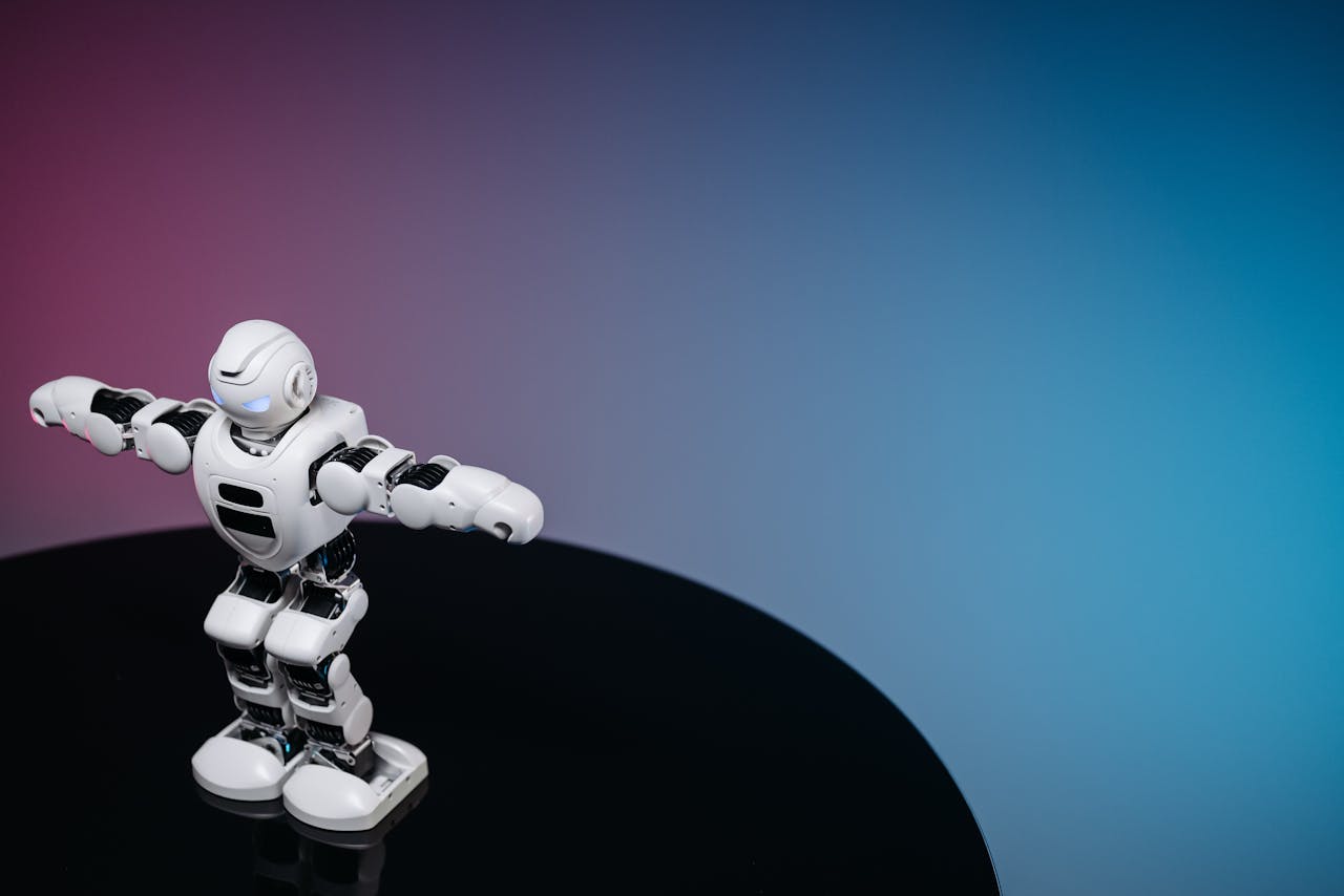 A white humanoid toy robot standing on a reflective black surface in a studio setting with a blue and pink gradient background.